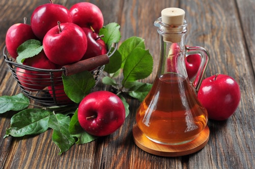 A bowl of red apples and a bottle of apple cider vinegar on a wooden table.