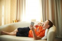 A young boy is lying on the couch playing with the phone in the room in an orange shirt is eating ice cream. Childhood inactivity is now a public health hazard. Motivate your kids to put away technology and get off the couch.