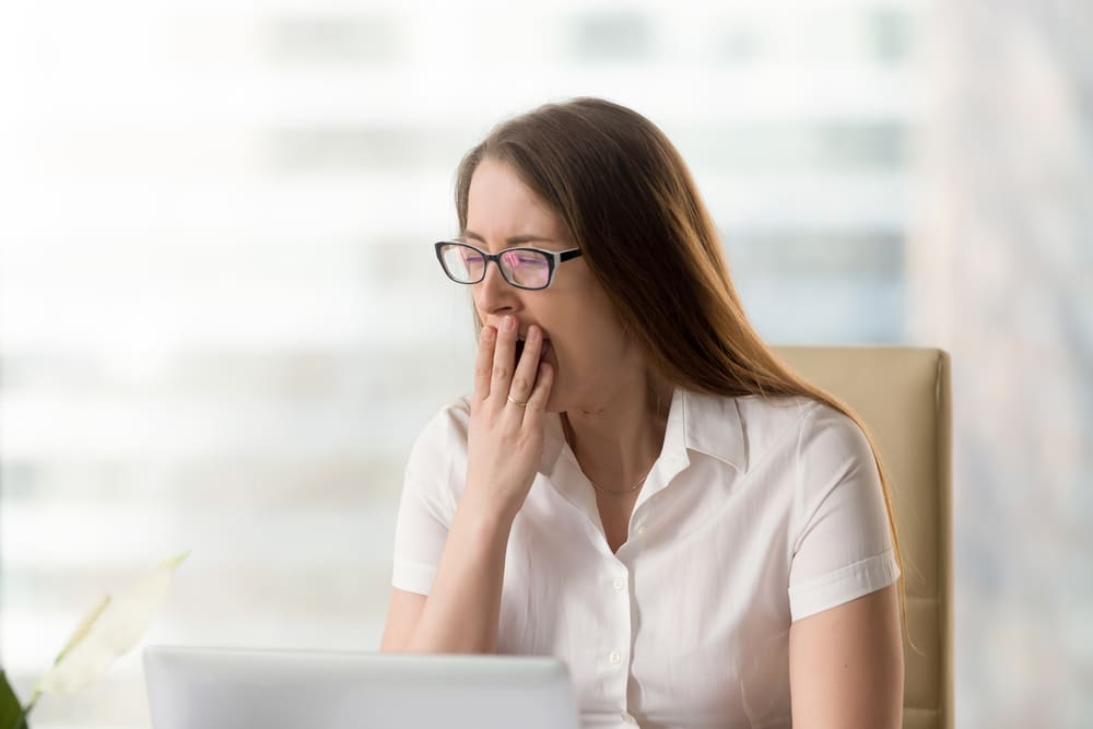 A woman at work yawning while sitting in her chair at her desk with her computer. Lear about 6 habits that can make you feel tired.