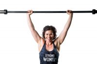 A happy and confident, muscular female trainer easily holds an unloaded barbell over her head getting ready to start her hypertrophy training