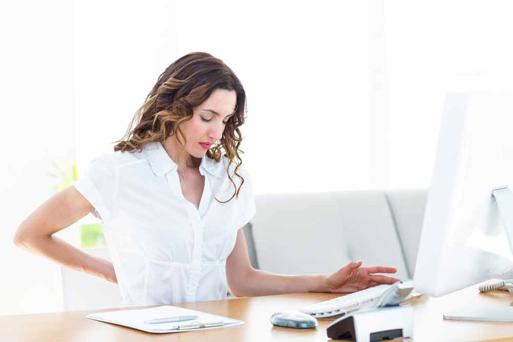 image of female worker sitting having back problems. When you sit too much lower back pain can be an unfortunate result.