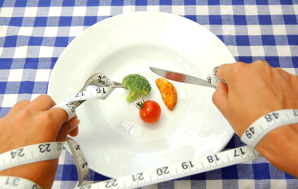 Tape measure wrapped around hands over a dinner plate containing an extremely small amount of food holding a knife and fork illustrating that calorie restriction doesn't work