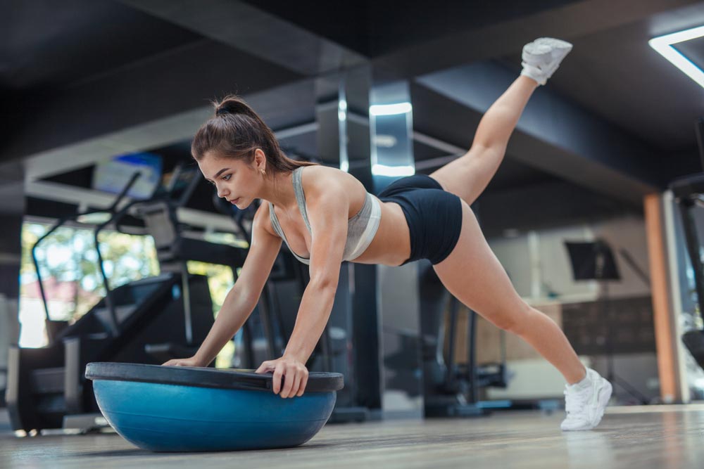 Young woman leaning on bosu ball and lifting legs with great concentration as she works on her balance skills.