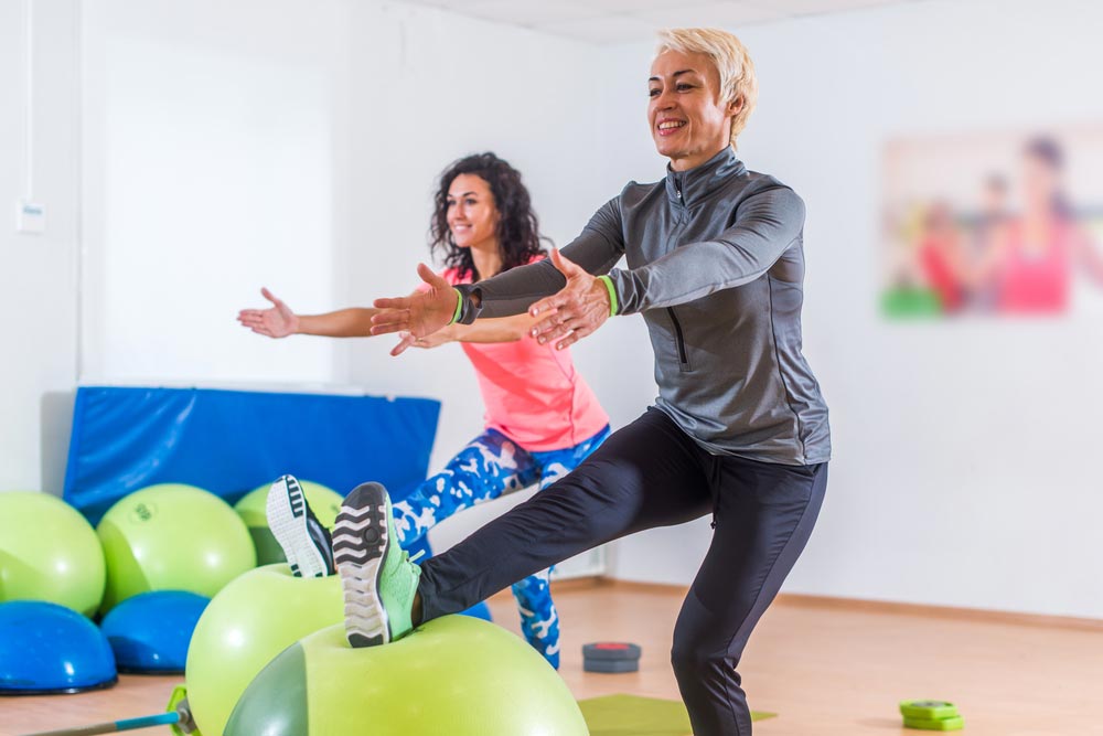 Group of active cheerful sporty women doing single-leg squats with balance ball training indoors in gym.